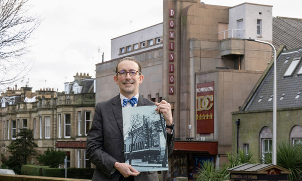 A man smiles at the camera whilst holding a large black and white photo of an art deco cinema building. He wear a grey suit, white shirt, blue striped bowtie and glasses. The cinema from the photo is in the background behind him.
