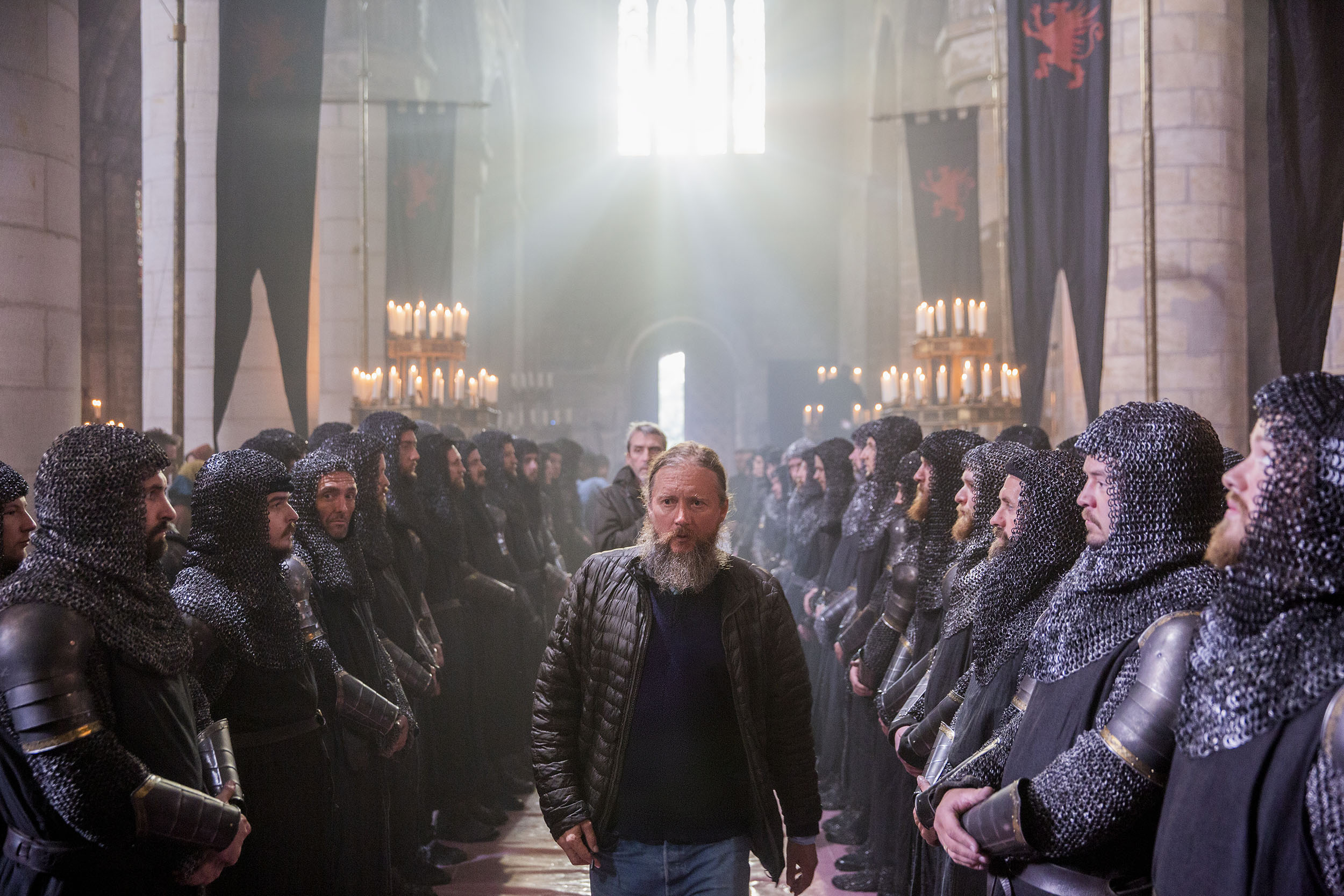 A modern‑dressed film director walks between two lines of armoured knights inside a grand candlelit hall during filming.