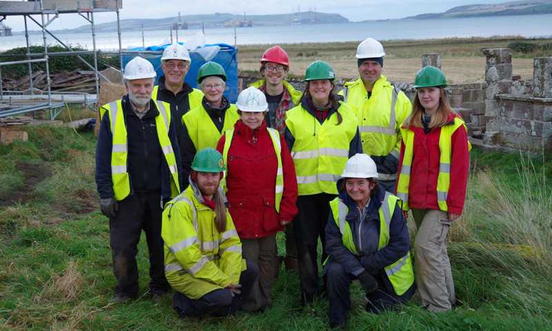 A photograph of a group of people wearing hard hats and high visibility jackets standing by scaffolding in a ruined building, smiling at the camera.