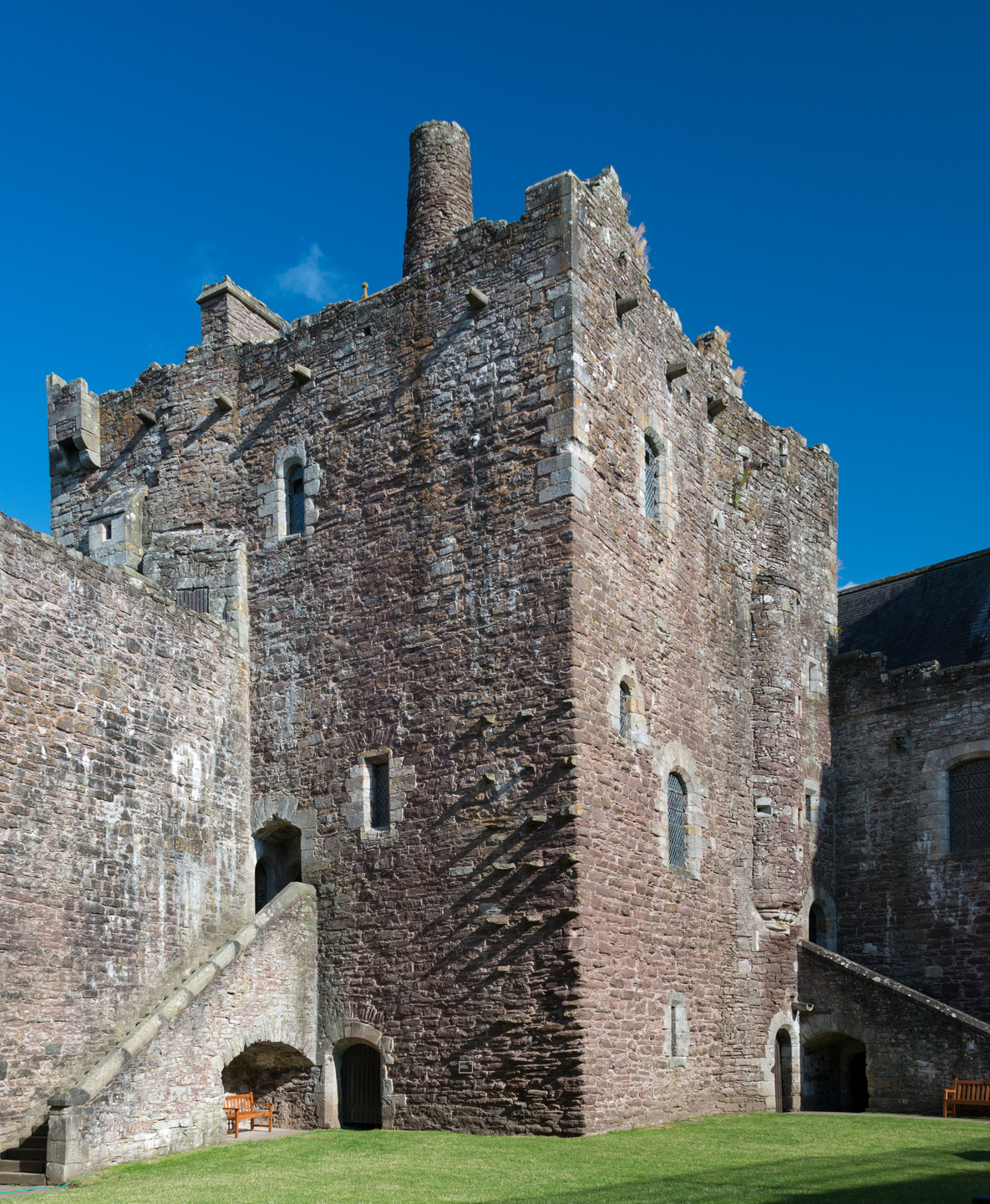 The side of a historic stone building, there are stairs on either side of it. There are square and arched windows on the building. It sits on a grass landscape.