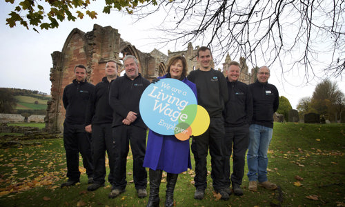 Cabinet Secretary Fiona Hyslop holding a sign that says "We are a Living Wage Employer", with Monument Conservation Team at Melrose Abbey 