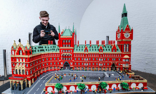 man uses phone to take a picture of large lego model of a red building with a clock tower