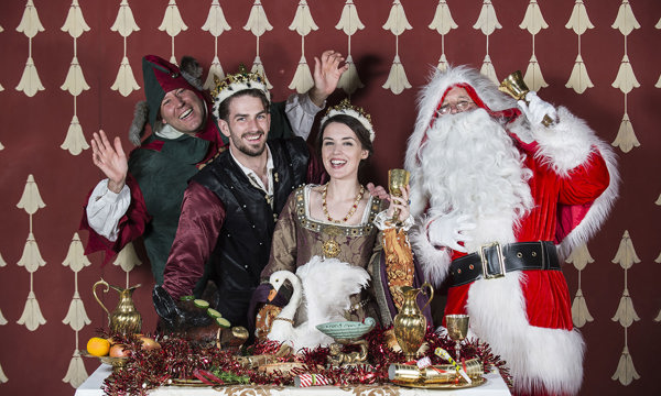 four smiling costumed performers stand in a row behind a table covered in festive props - a jester, a male courtier, a renaissance queen and father christmas