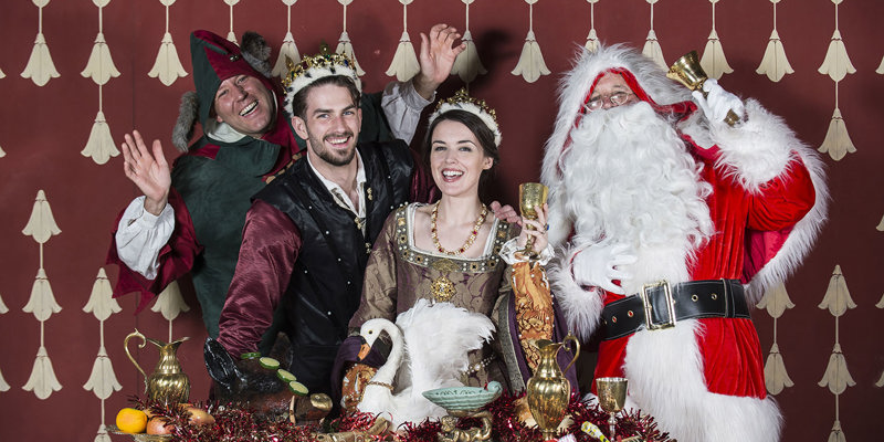 four smiling costumed performers stand in a row behind a table covered in festive props - a jester, a male courtier, a renaissance queen and father christmas