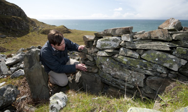 A man crouches next to a ruined stone wall with grass and a large body of water behind him