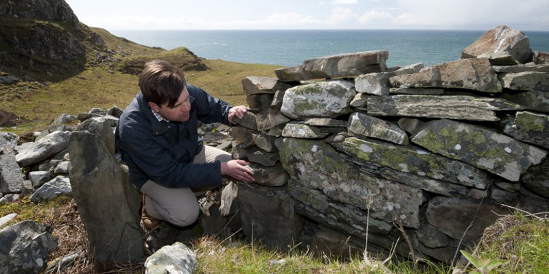 A man crouches next to a ruined stone wall with grass and a large body of water behind him