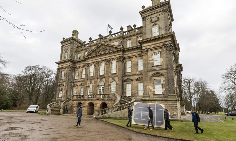 five people carry a large painting towards the stairs of an elaborate mansion house whilst a man directs them