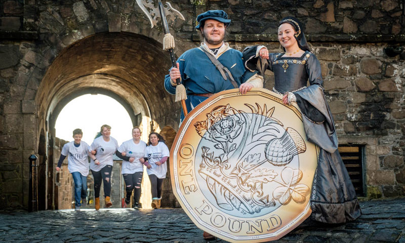 Two re-enactors holding a giant pound coin, with four children behind them running out of a castle