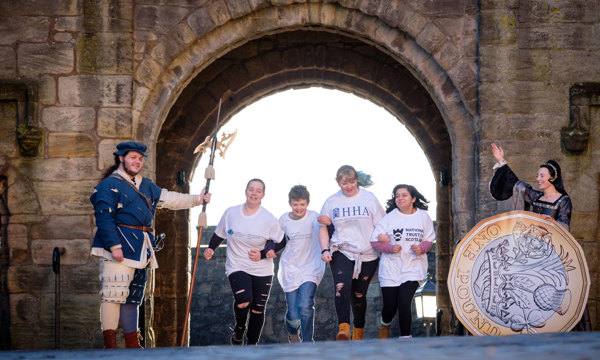 Four children run through a castle archway with one re-enactor at either side, one of which has a giant pound coin