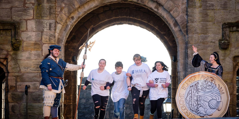 Four children run through a castle archway with one re-enactor at either side, one of which has a giant pound coin