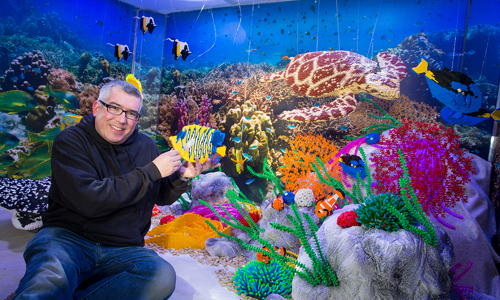 man sits in front of an underwater backdrop with model fish around him