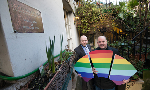 two men stand outside old building holding rainbow keystone 