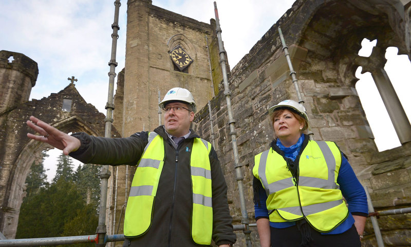 A photograph of a man and a woman wearing white hard hats and yellow high visibility jackets, standing in front of a historic church building
