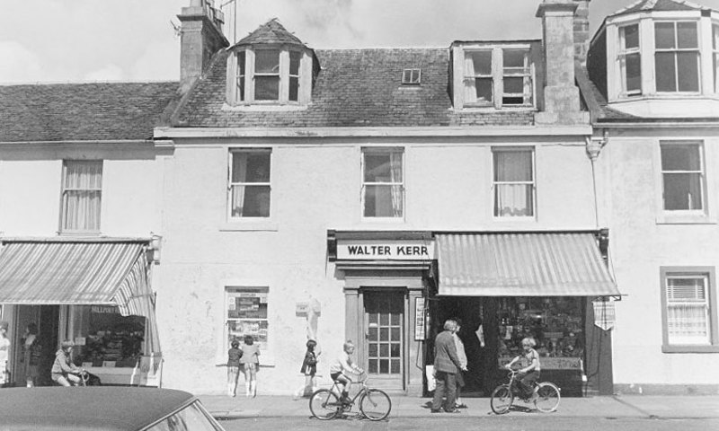 A black and white photograph of a building with a shop front. Children on bicycles and and people looking in windows are walking past.