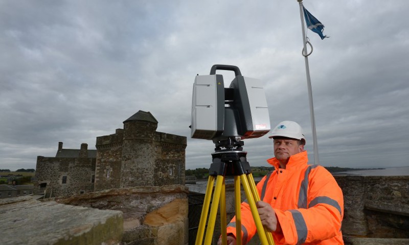 A photograph of a man in an orange high visibility jacket looking at a laser scanner in front of a castle