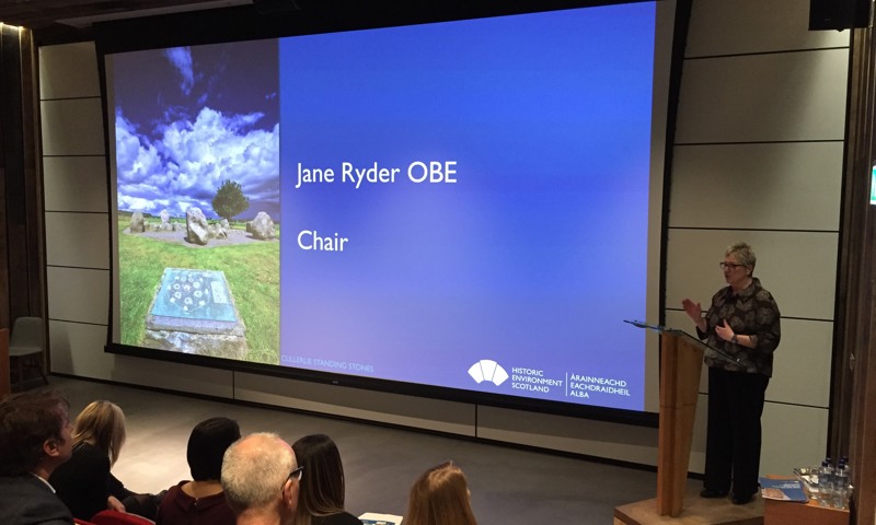 A photograph of a woman speaking to an auditorium. The name on the presentation reads "Jane Ryder OBE"