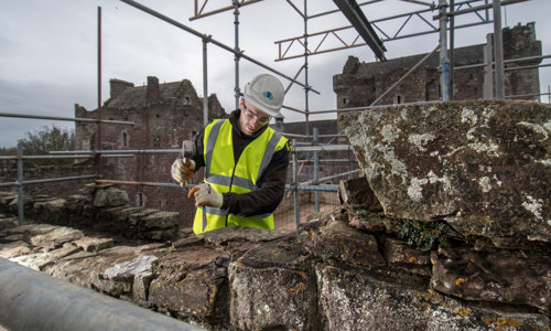 A photograph of a man in a yellow high visibility jacket and hard hat chiseling at stone on the top of a castle 