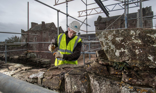 A photograph of a man in a yellow high visibility jacket and hard hat chiseling at stone on the top of a castle 