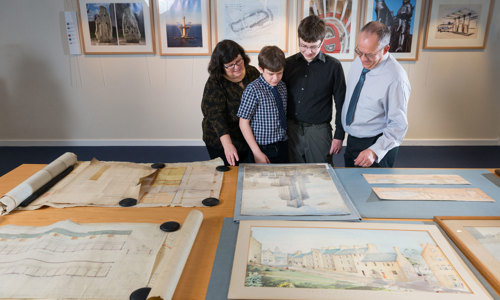 A photograph of a family looking at drawings and paintings of architecture laid out on a table