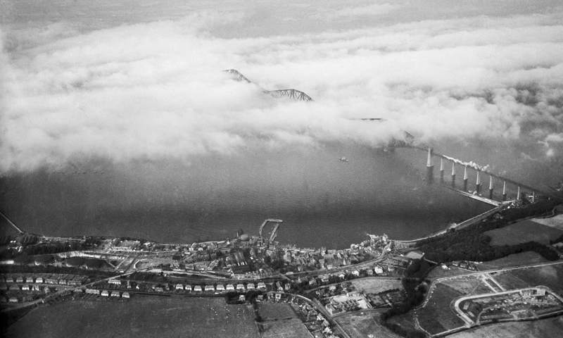 A black and white photograph taken from the air of a bridge disappearing into clouds and a town on the edge of the water.