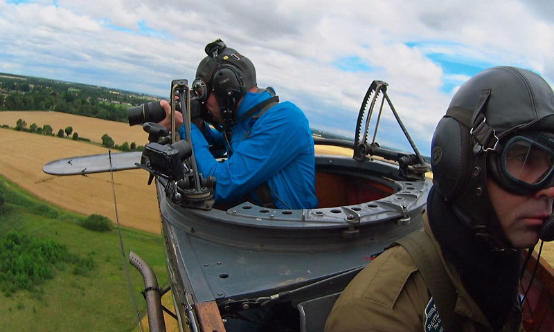 A photograph of two men in an open-top plane. One man is wearing a flying helmet and goggles. The other is wearing a blue jacket and is looking through a camera down at the land below.