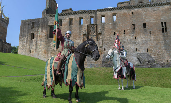 Two men in medieval knight costumes on horseback pose holding flags above their heads in front of a large stone castle.