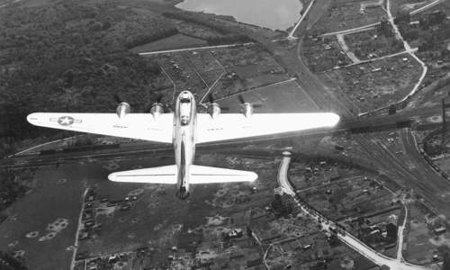 A black and white photograph take from above of a plane flying over a bombed landscape