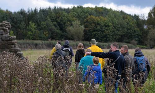 A photograph of a group of people walking through a field beside a ruined historic building towards a wood