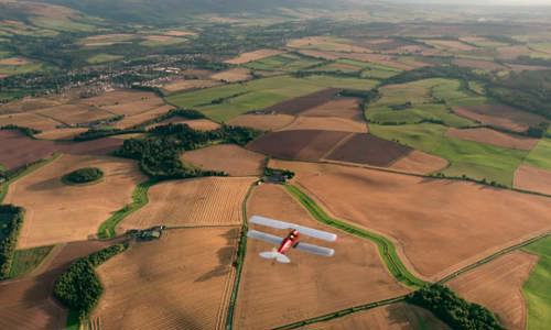 An aerial photograph of a red and white plane flying over fields of gold and green