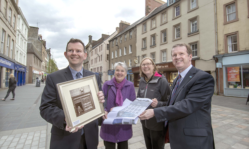 four people stand in a row holding plans and sepia photo of buildings around them