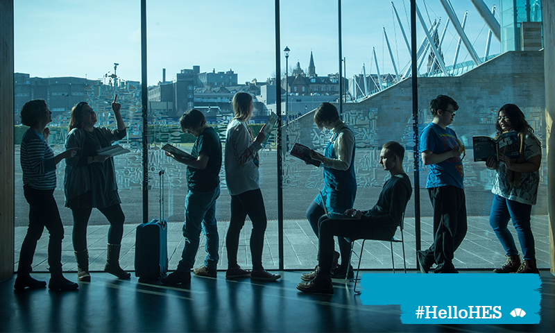 A photograph of eight people standing and sitting beside a window. They are reading and looking up at the building. Outside, there is a city view.