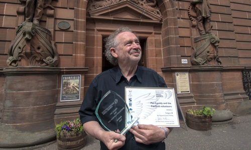 A man stands outside a red sandstone building holding an award and certificate his organisation received for a building restoration project
