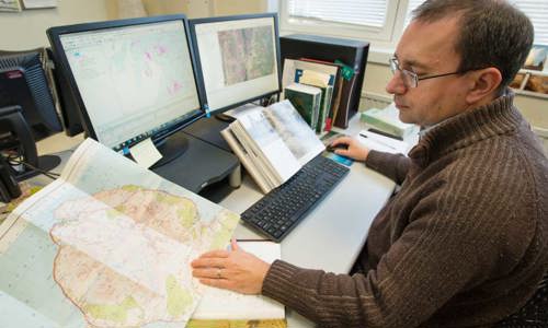 A photograph of a man wearing glasses, looking at a map with a digital version of the map on the computer in front of him