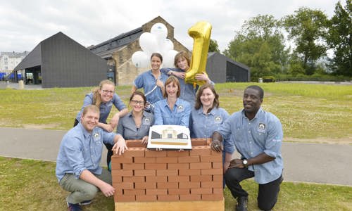 Members of staff stand outside the Engine Shed in front of a fake brick wall, smiling, wearing Engine Shed shirts, holding up balloons 