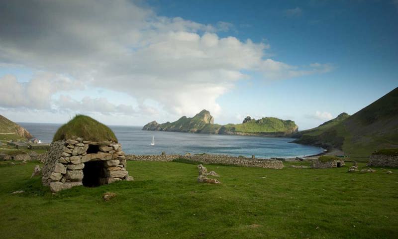 A small stone hut by the sea with green hills nearby.