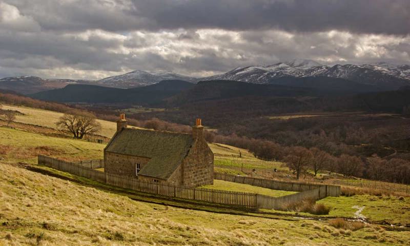 A small, isolated cottage on a hill with large snow-capped mountains in the distance.