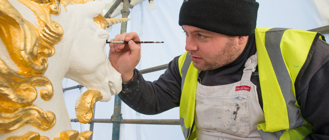 A man in a high visibility jacket and a beanie hat painting a carved unicorn head.