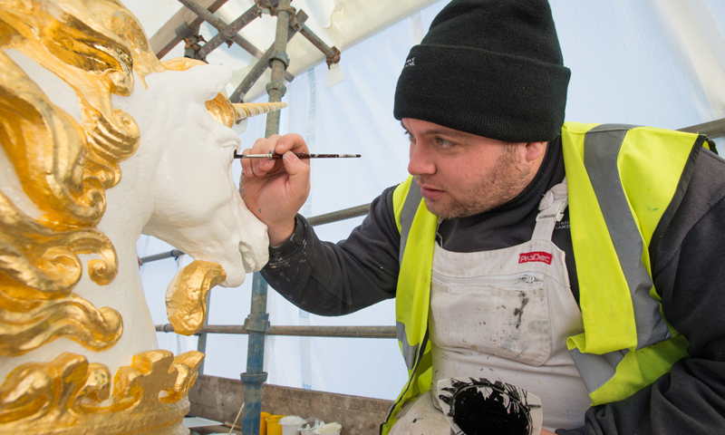 A man in a high visibility jacket and a beanie hat painting a carved unicorn head.