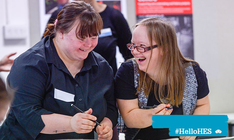 A photograph of two women with disabilities holding pens and laughing together.