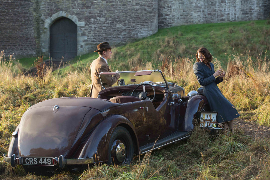 Two Outlander cast members standing next to an old fashioned open top car with the entrance door of Doune Castle in the background
