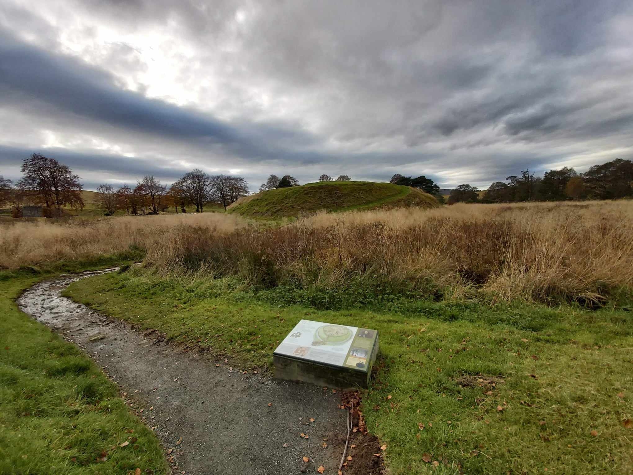 Winding gravel path with an information plaque in the foreground, leading to the Lumphanan earthwork mound surrounded by tall dry grass and trees under a cloudy sky.