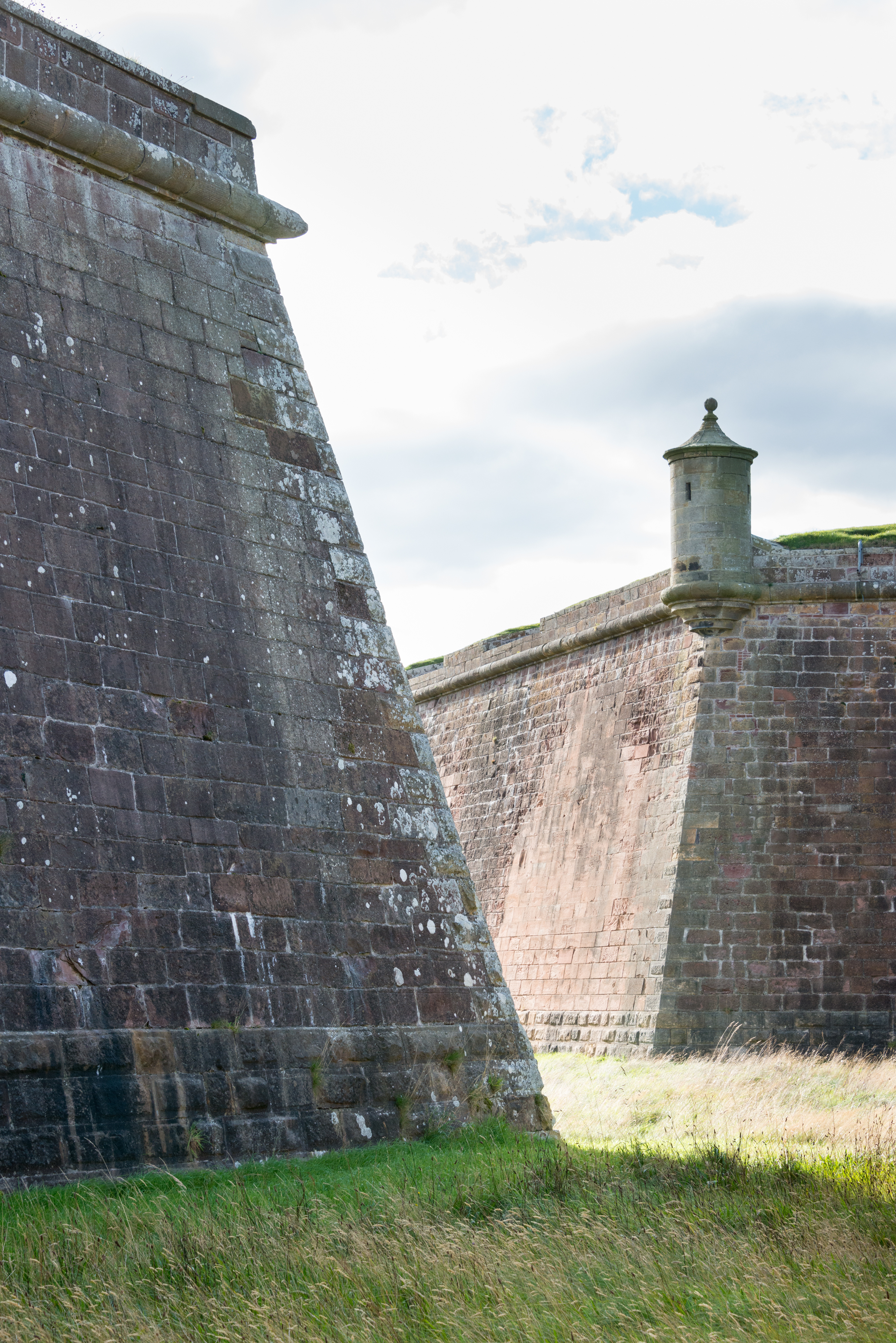 Close-up view of two massive, angled stone bastions at Fort George, with weathered stonework and long grass at their base.