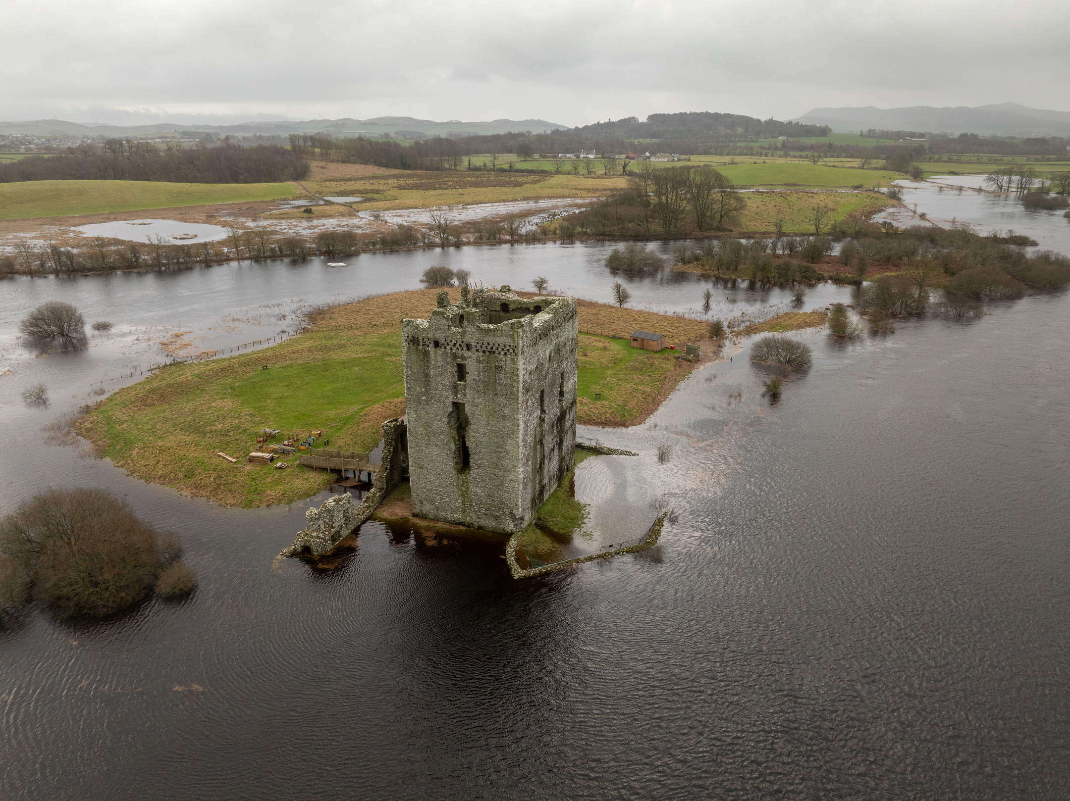 Aerial view of Threave Castle standing on a small grassy island, surrounded by dark floodwater that has overflowed into the surrounding fields and trees.