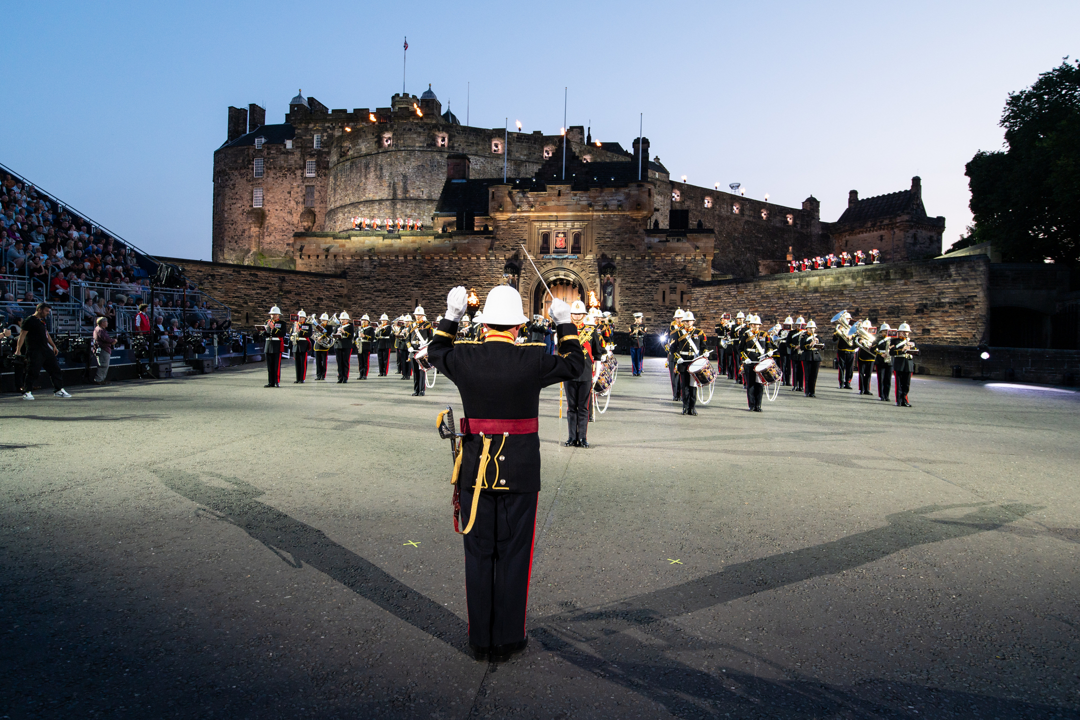 Military band performing in formation on the castle esplanade at dusk, with Edinburgh Castle lit in the background and audience seating to the side.