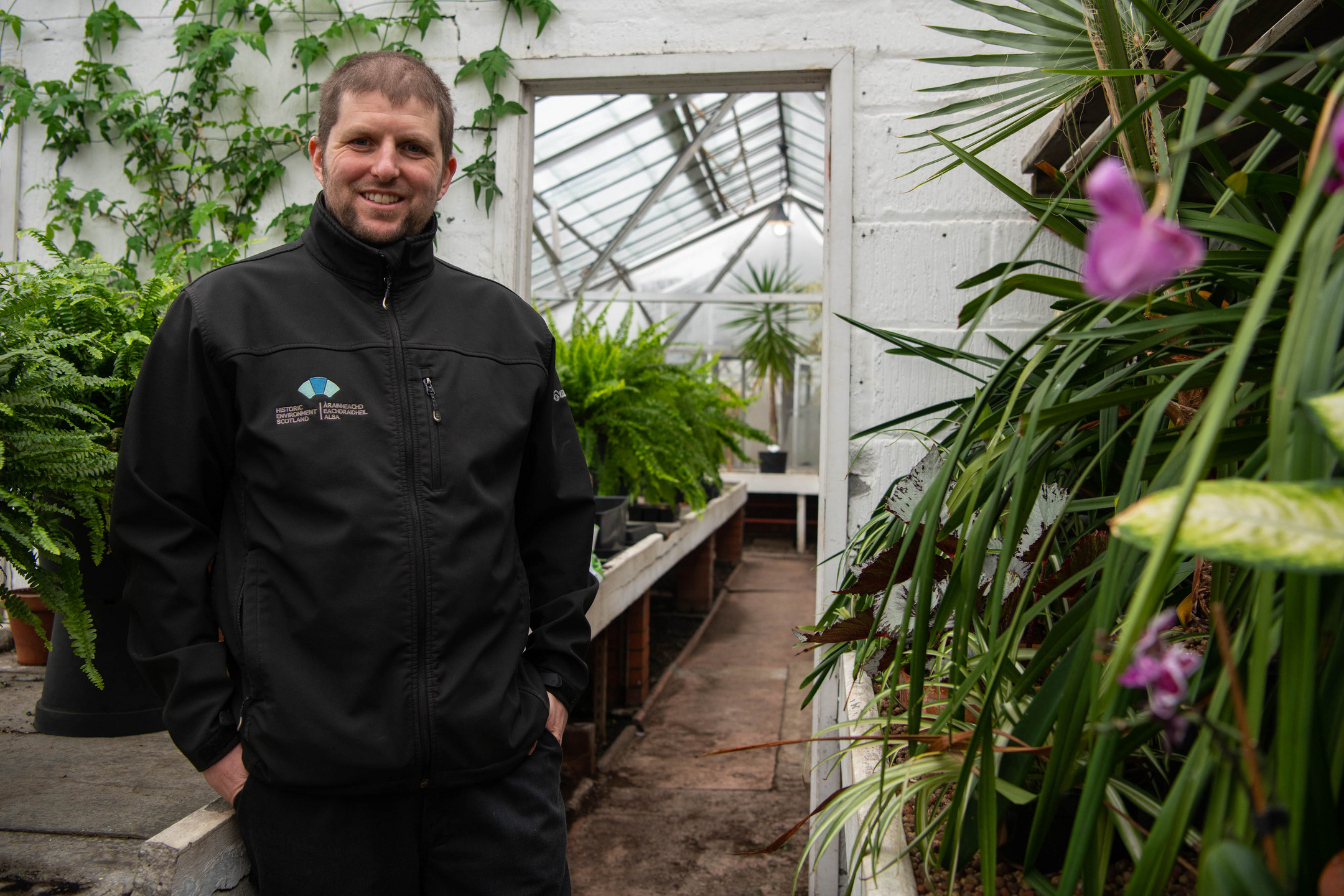 Portrait of a gardener surrounded by plants in a greenhouse. The man is wearing Historic Environment Scotland clothing.