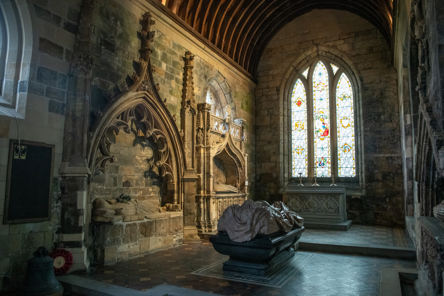 Carved stone tombs and monuments along the church walls, with a large stained-glass window at the back and a stone effigy in the centre.