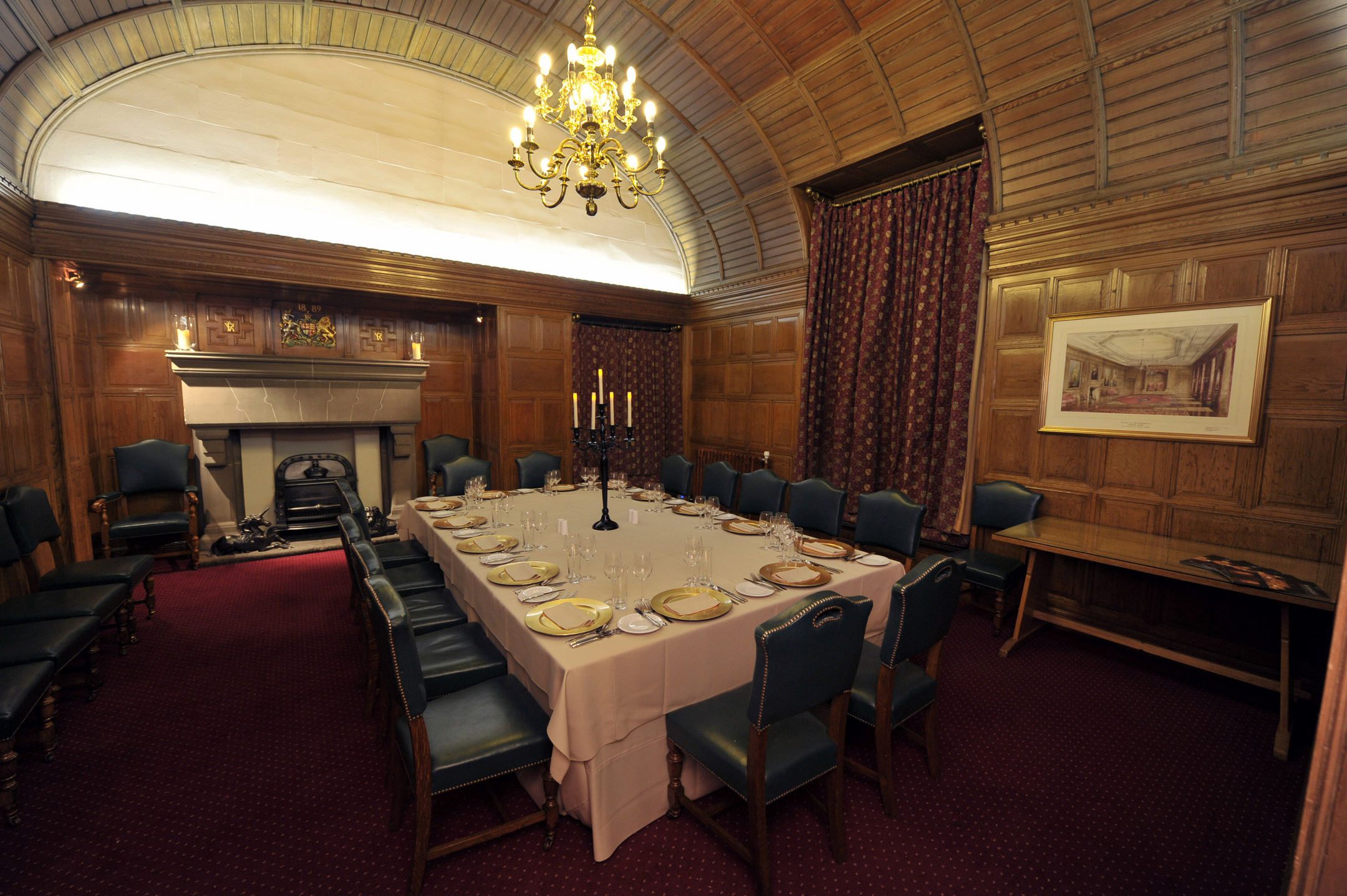 A large dining room table is set in the centre of a historic wood paneled room. There is a large fireplace at one end, and a chandelier lights the room.