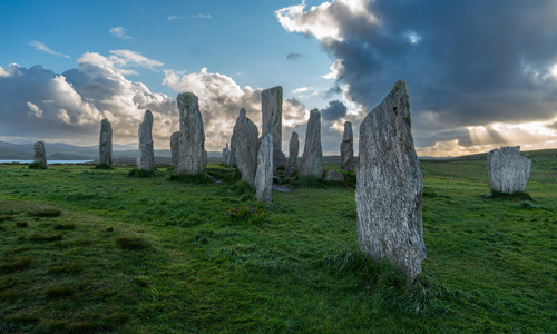 The Calanais Standing Stones pictured against a blue sky with large, ominous clouds. The clouds are mostly darks, but dwindling sunlight breaks through in the distance, and highlights the edges.