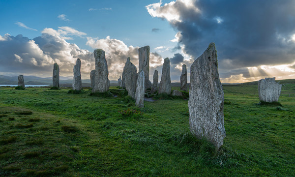 The Calanais Standing Stones pictured against a blue sky with large, ominous clouds. The clouds are mostly darks, but dwindling sunlight breaks through in the distance, and highlights the edges.