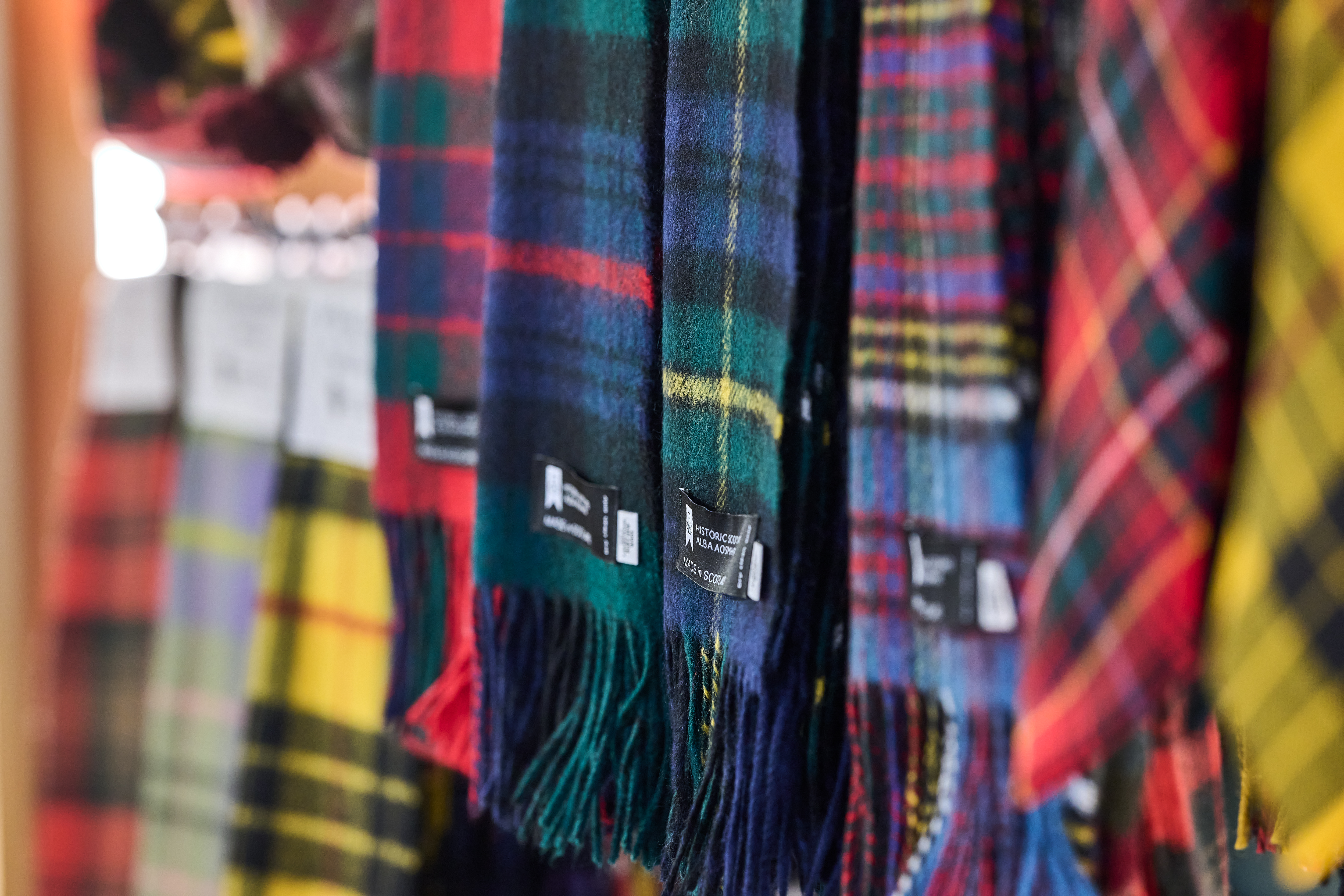 A rack of colourful tartan scarves hanging in a row, showing patterns in greens, reds, blues, and yellows. Product labels are visible along the edges.